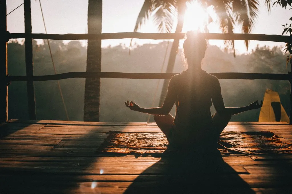 Silhouette of a person meditating in lotus position on a wooden deck at sunrise, framed by palm trees during the Bali writers retreat