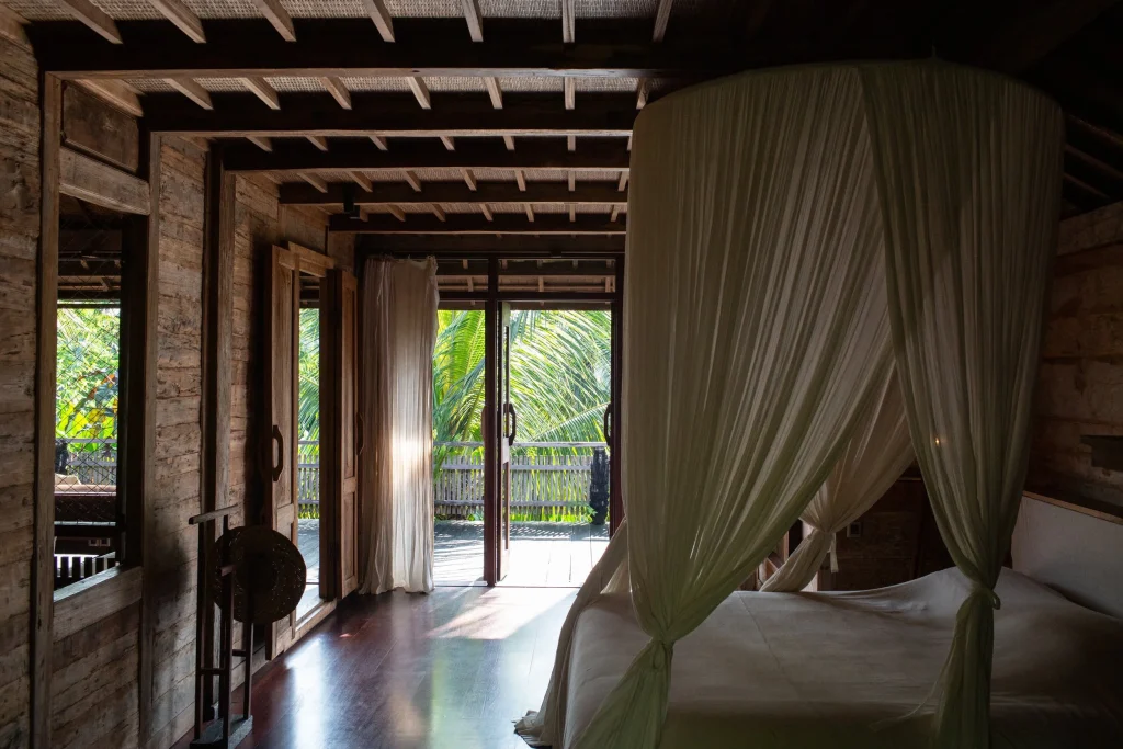 Wooden bungalow bedroom with a four-poster bed draped in sheer white curtains, opening onto a terrace with palm trees at the Bali eco-lodge