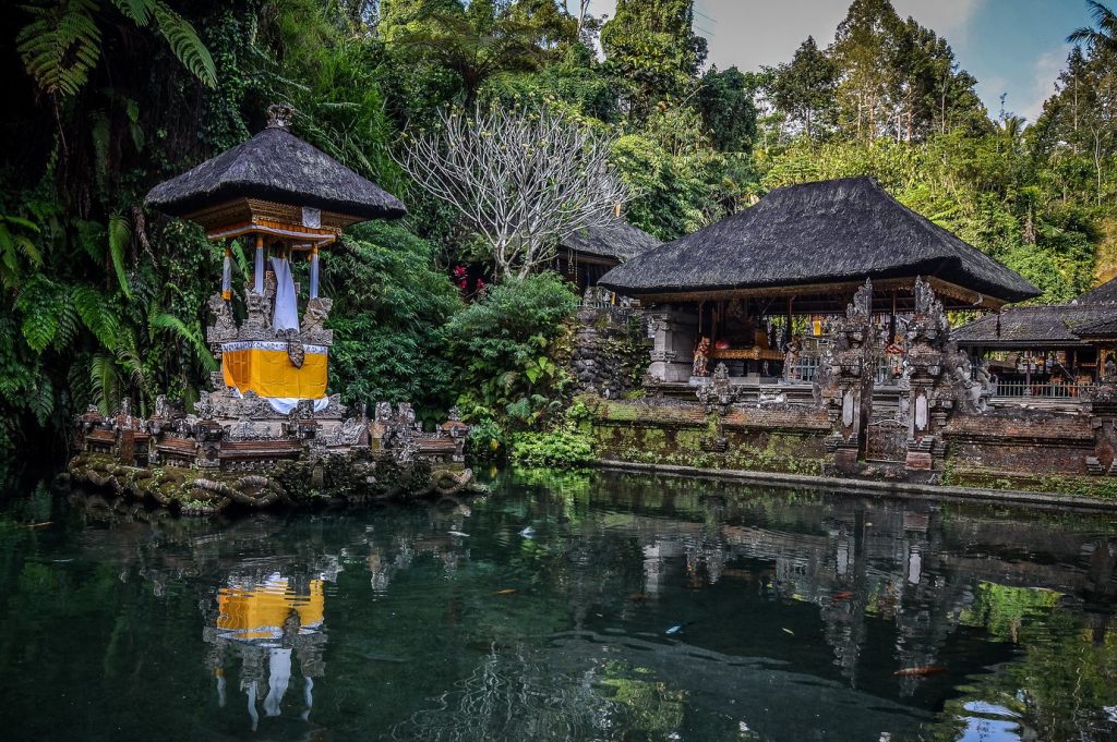 A traditional Balinese temple with tiered thatched roofs and shrines draped in ceremonial cloth, beside a tranquil fish pond surrounded by tropical forest