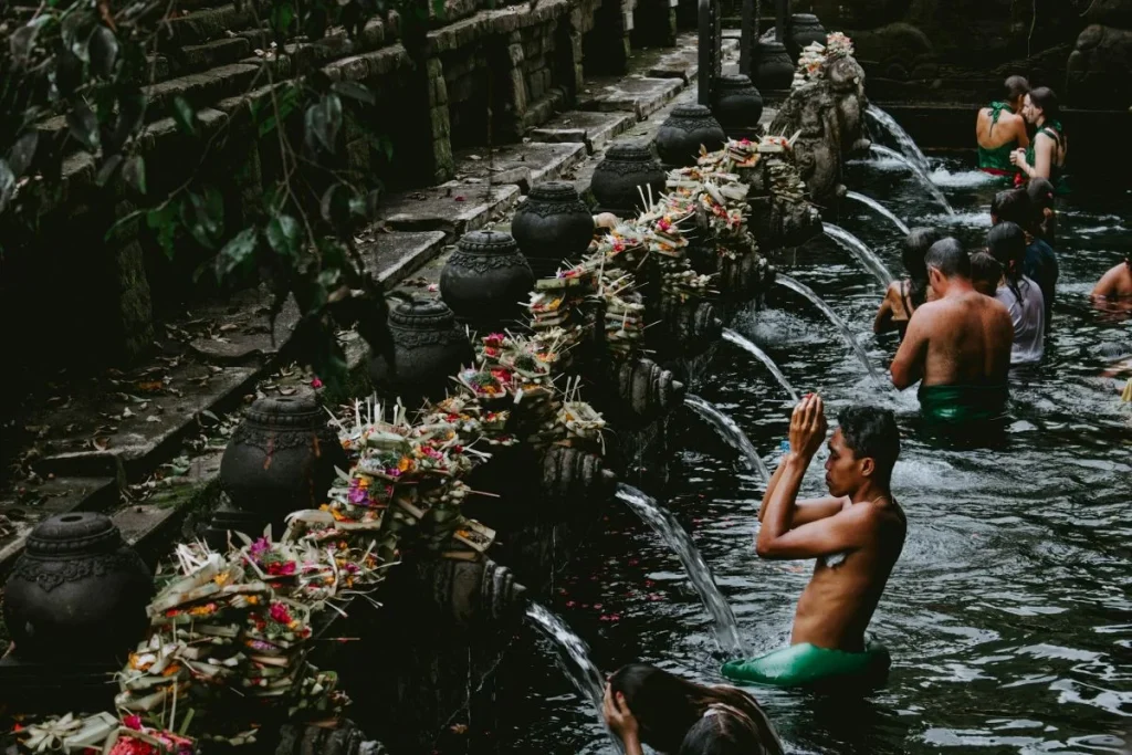 Visitors performing a traditional Balinese water purification ritual at a sacred spring temple in Bali, bathing beneath carved stone spouts lined with floral offerings
