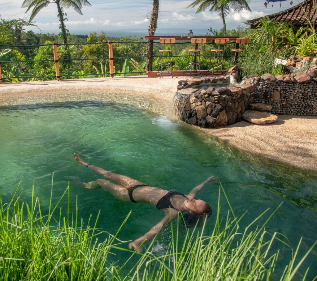 A guest floating in a natural stone-edged pool with a small waterfall feature and mountain views at the Bali retreat
